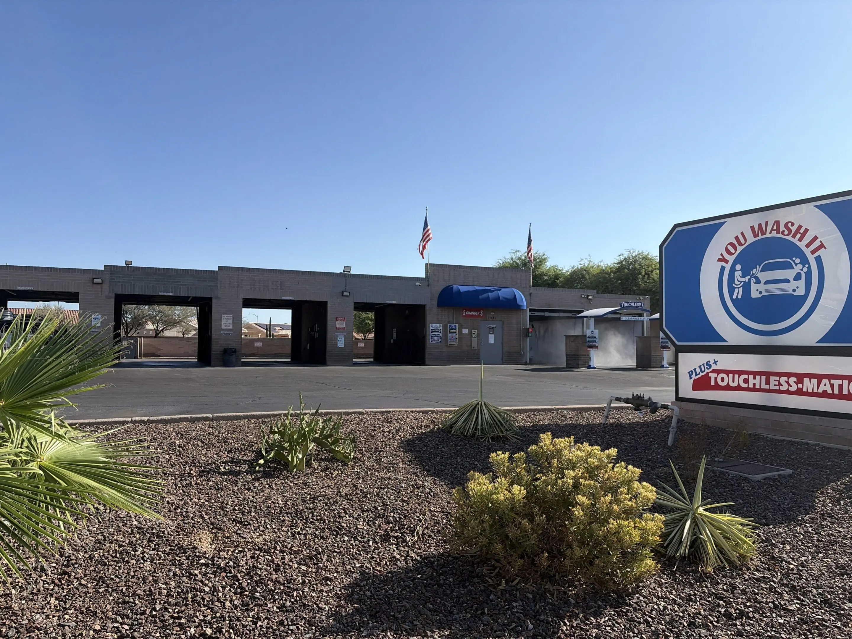 Building with a sign that reads "You Wash It" in front of it, surrounded by plants, stones, a road